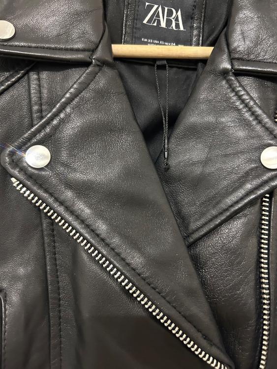 Black leather jacket with a zip and silver buttons, hanging on a wooden hanger. Visible label: ZARA.