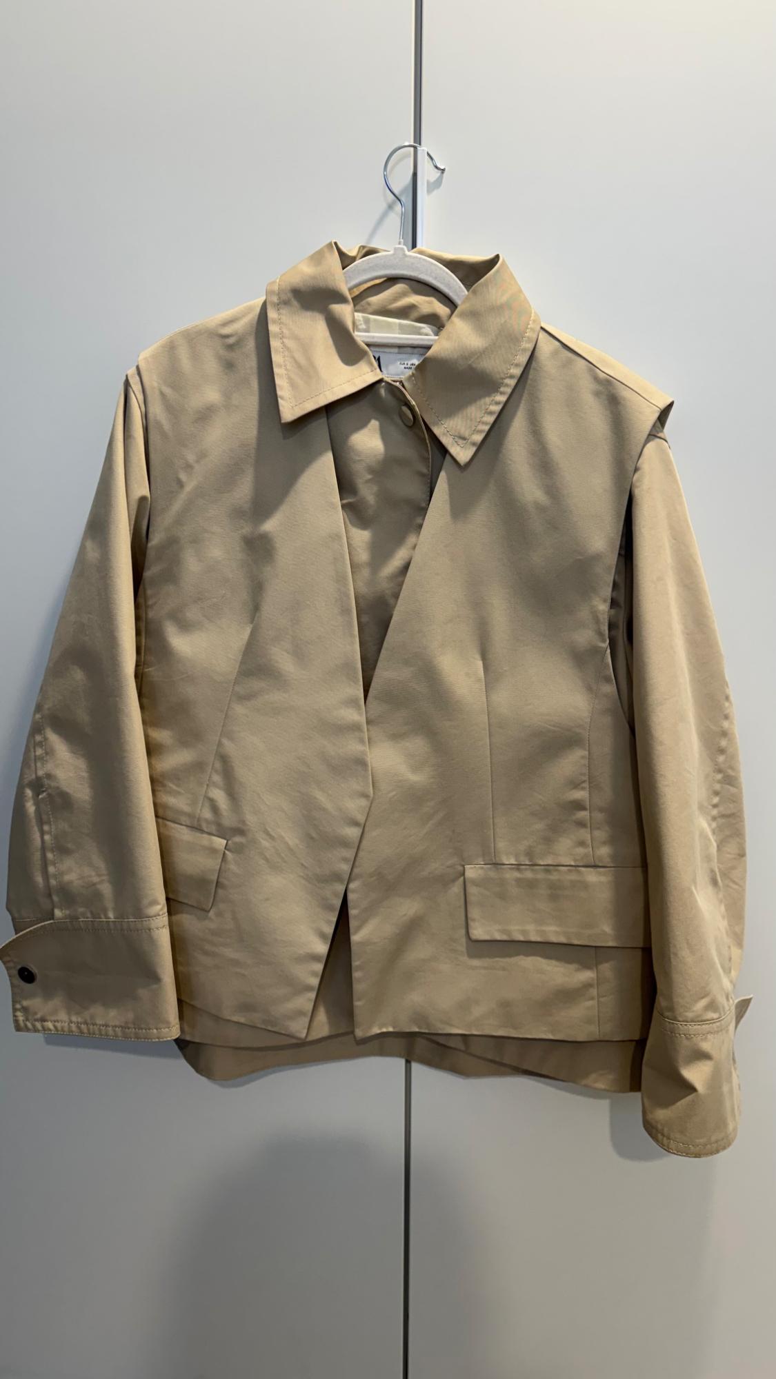 Beige jacket hanging on a white hanger against a light background. Minimalist design with lapels and front pockets.