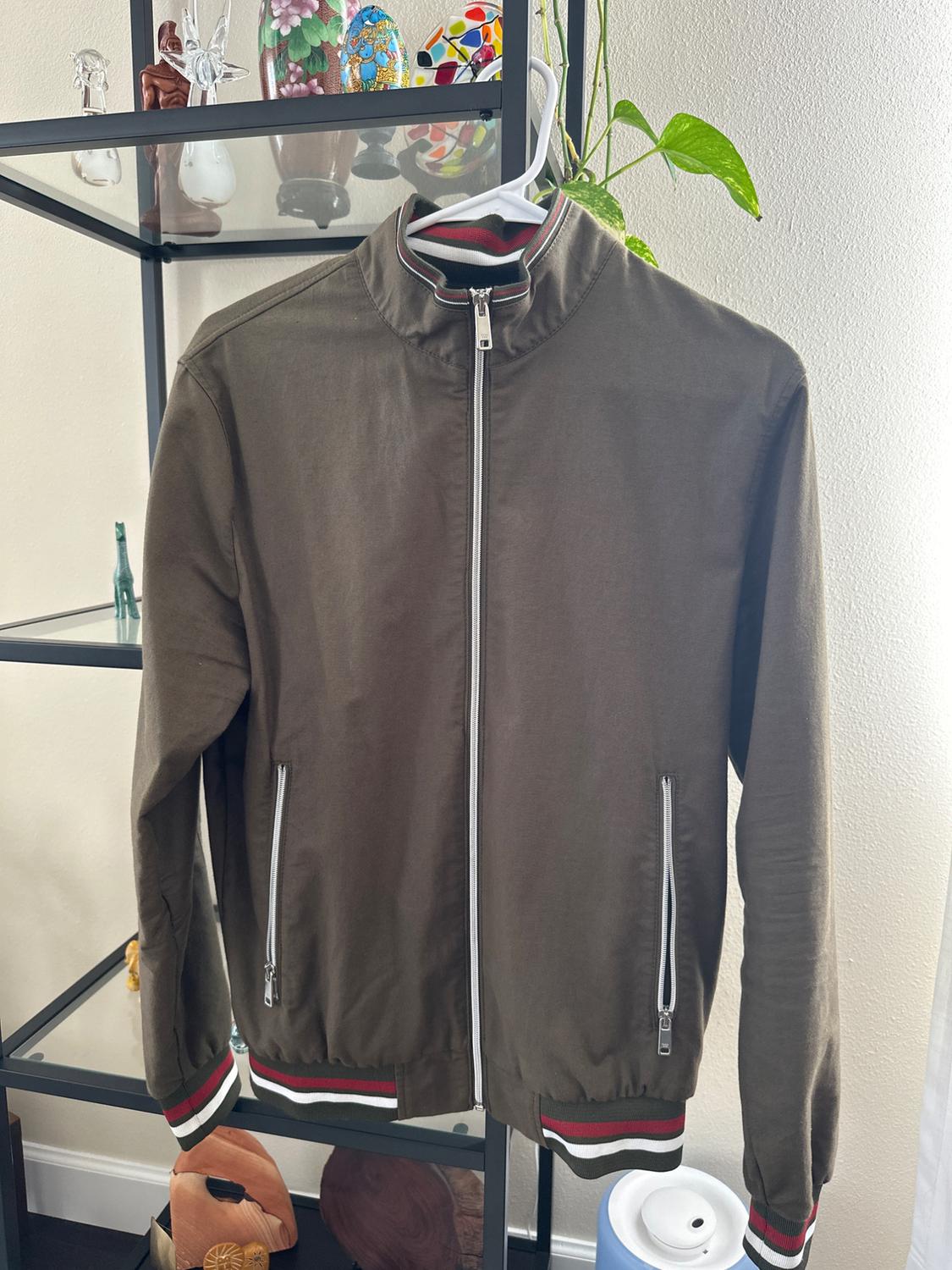 Olive green jacket with front fastening and zip pockets, detailing on cuffs and collar with red, white and black stripes, hanging on a coat hanger.