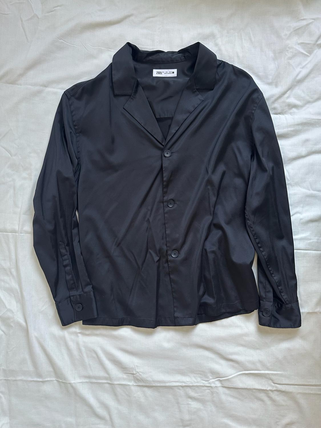 Black long-sleeved shirt with a lapel collar and buttons, spread out on a crumpled white sheet. Label visible with the text ZARA.