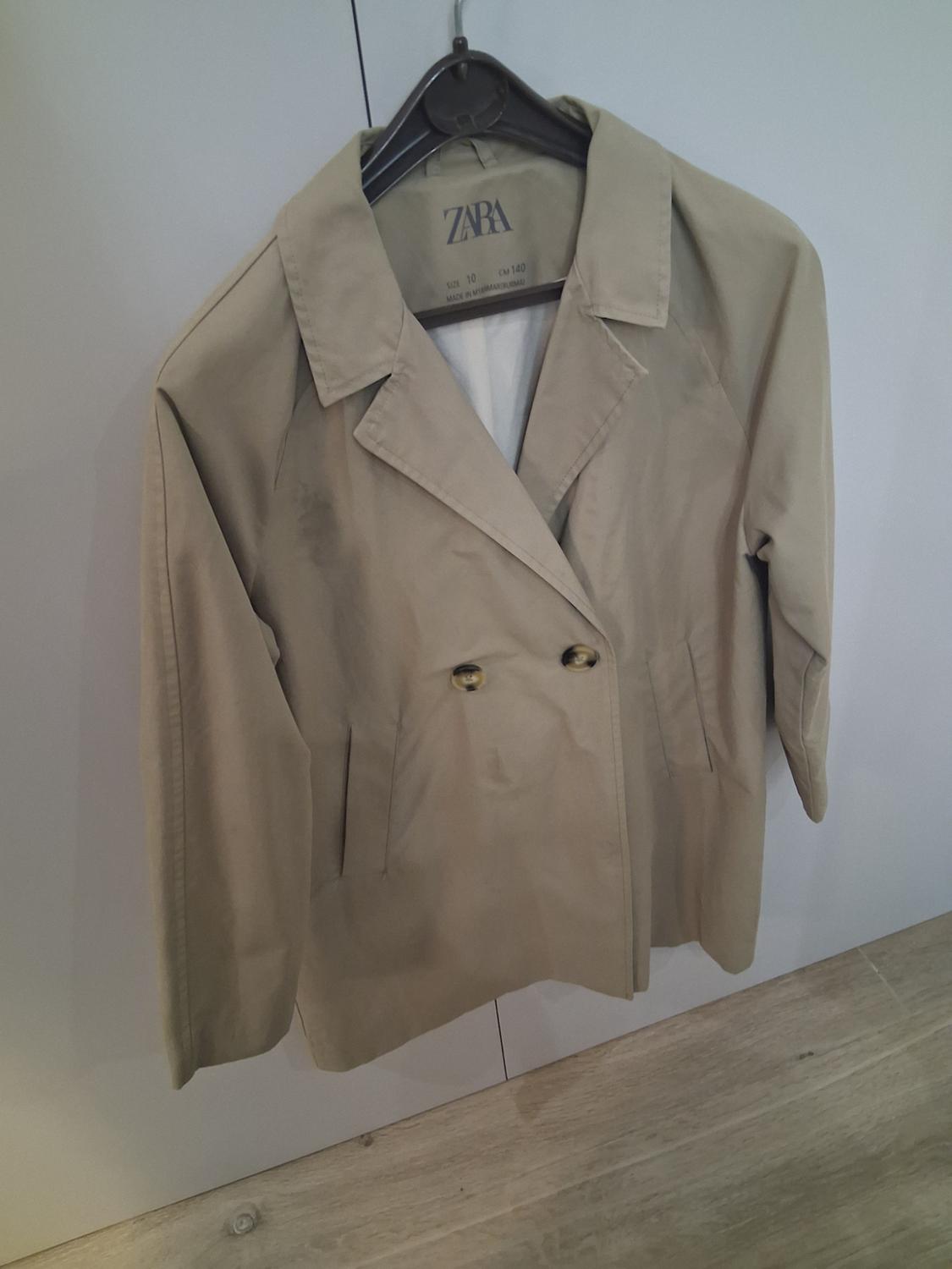 Beige double-breasted coat hanging on a black hanger. Visible label: "ZARA, UK 10, CM 140, MADE IN MOROCCO". White background.
