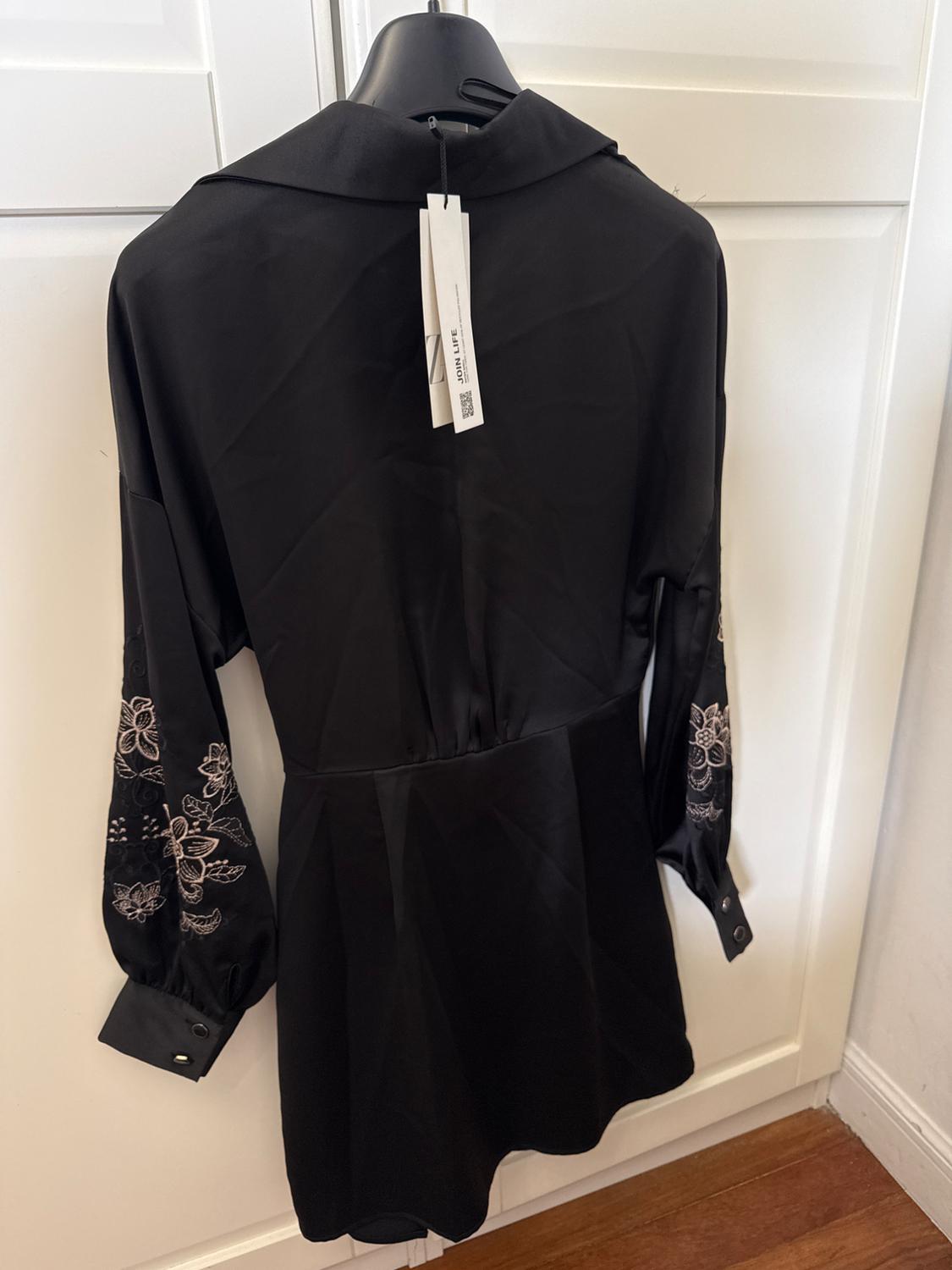 Black dress, viewed from behind, hanging on a white door. The floral embroidery on the sleeves and a 'JOIN LIFE' tag are prominent.