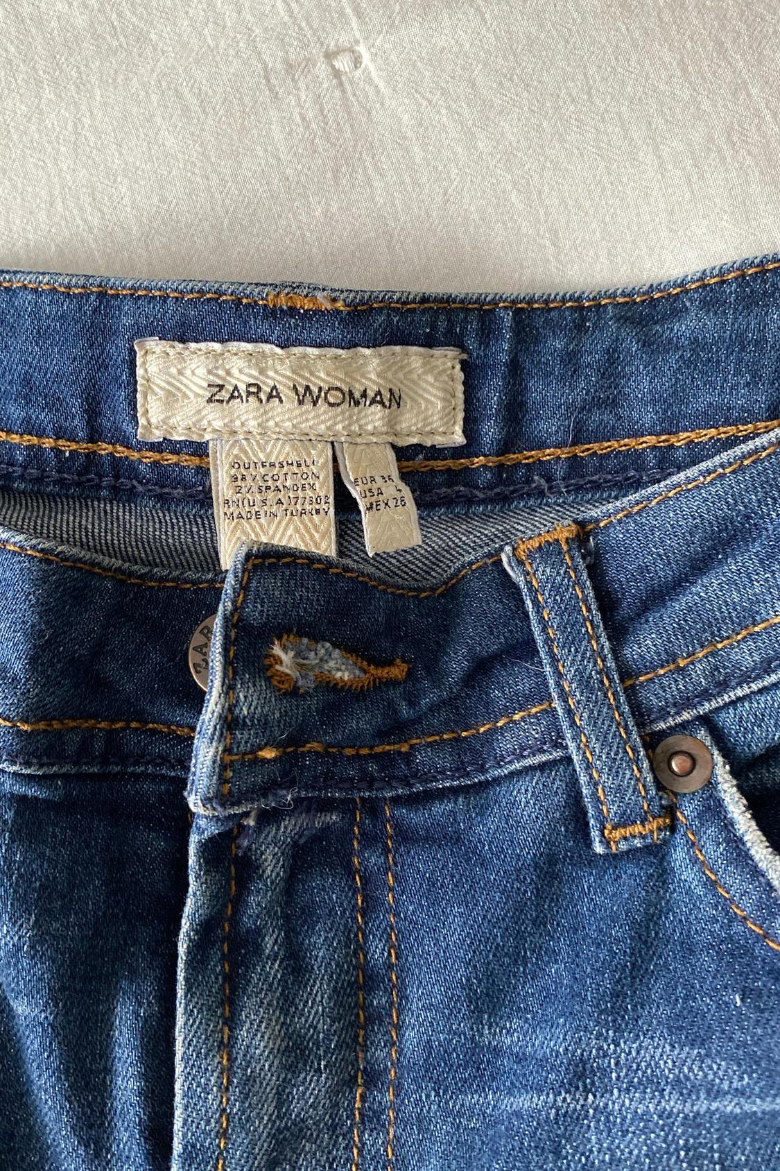 Detail of 'ZARA WOMAN' label on blue denim jeans with visible stitching and belt loops.