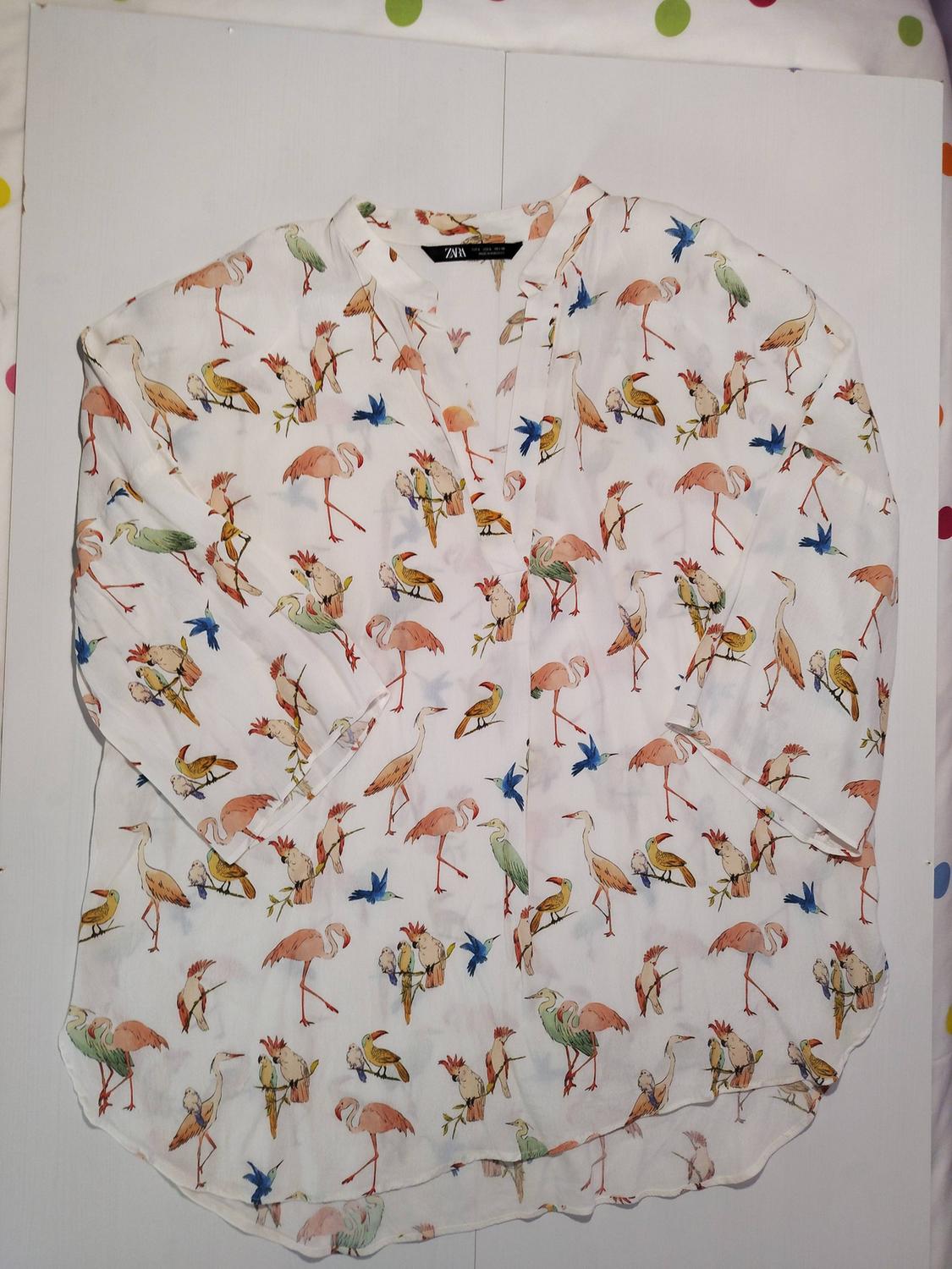 White blouse with a colourful bird print, including flamingos and toucans, on a white background. Visible label on the collar.