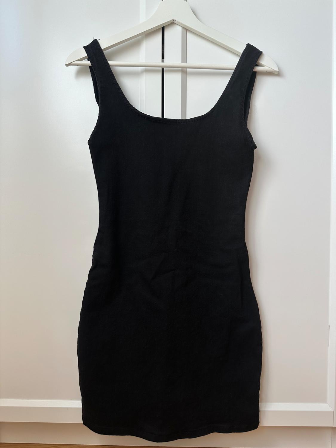 Black, fitted mini dress with wide straps, hanging on a white hanger in front of a white wardrobe. Minimalist style.