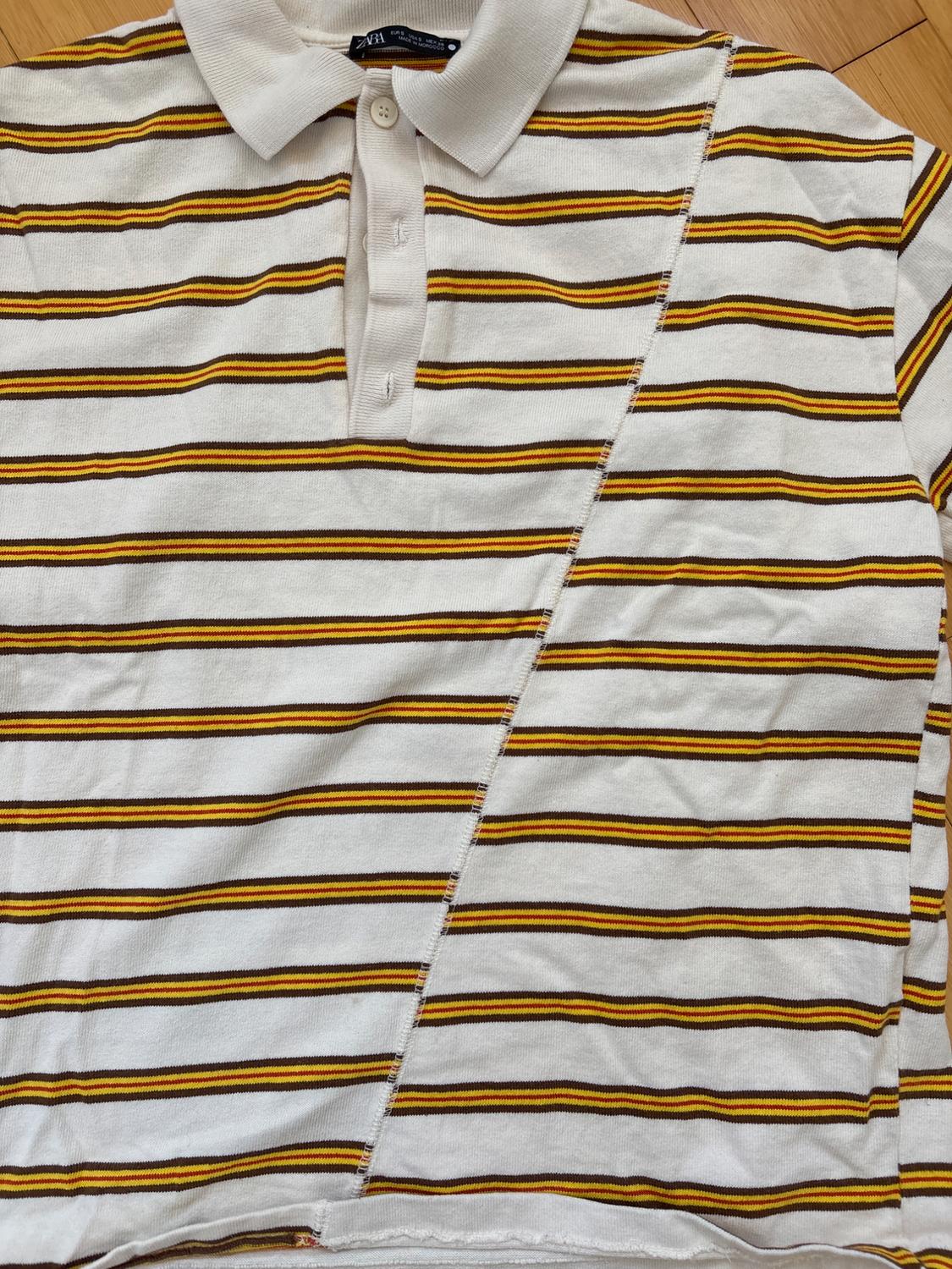 White, yellow, and black horizontal striped polo shirt, with an asymmetrical design and button-down collar on a light wood background.