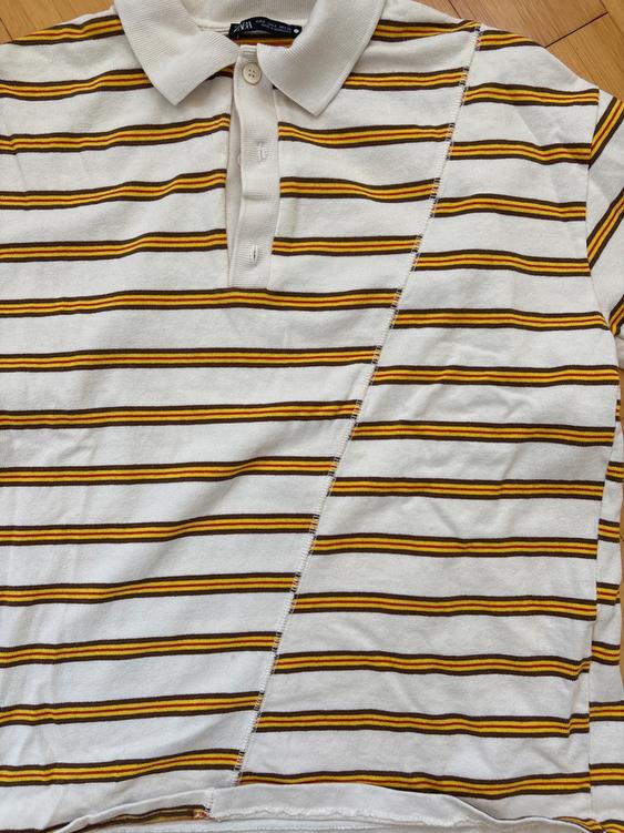 White, yellow, and black horizontal striped polo shirt, with an asymmetrical design and button-down collar on a light wood background.