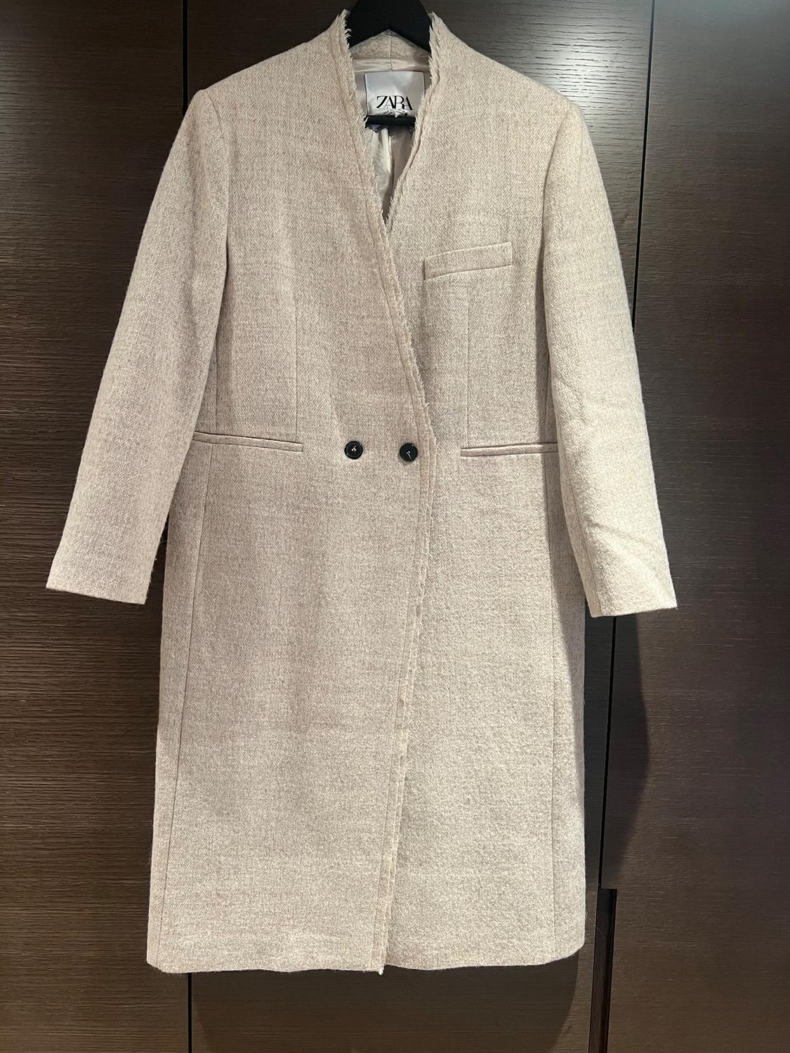 Beige, straight-cut double-breasted coat with a chest pocket, hanging on a hanger against a dark wood background.
