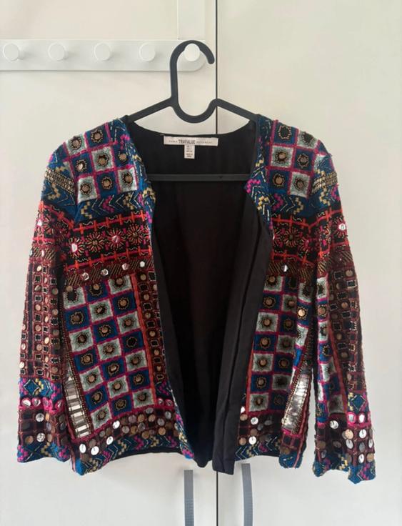 Jacket hanging on a hanger, with a colourful and geometric design, adorned with mirrors and embroidery. White background with hooks.