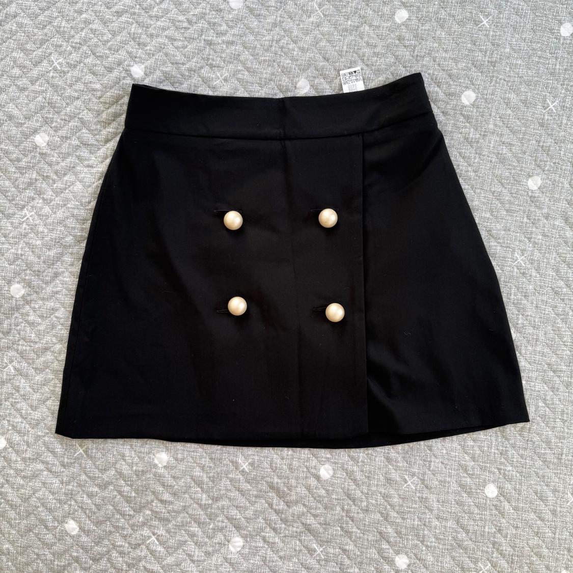Short black skirt with gold buttons in two rows, on a gray background with a diamond and white dot pattern. Label visible at the top.