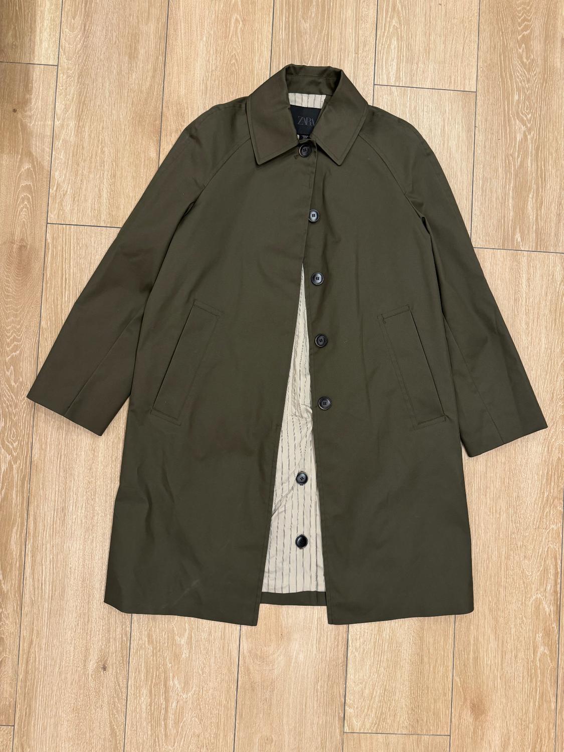 Olive green coat with black buttons and striped inner lining, laid out on a light wooden floor. Visible label on the collar.