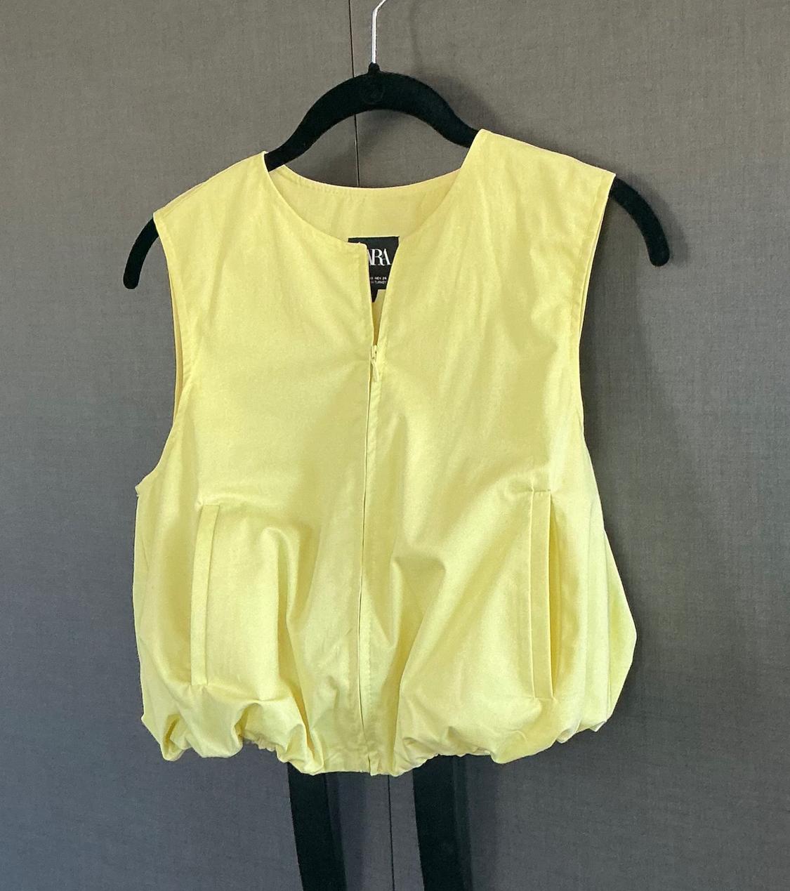 Yellow sleeveless top with pockets and a bubble hem, hanging on a black hanger against a dark background. ZARA text on the label.