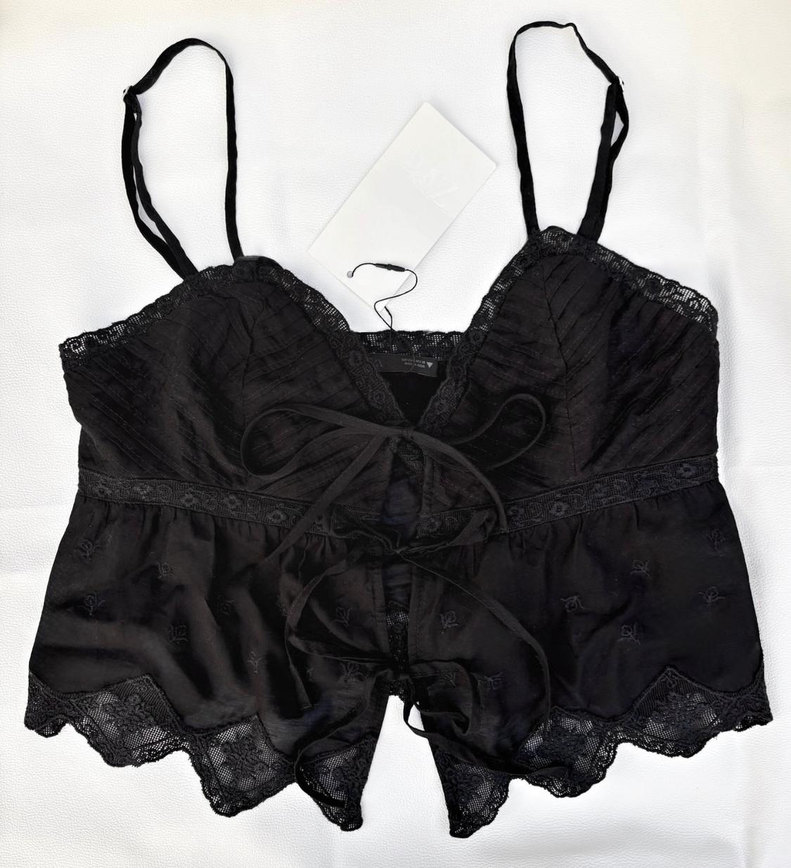 Black strappy top with lace detailing, subtle embroidery and front bows, presented on a white fabric background.