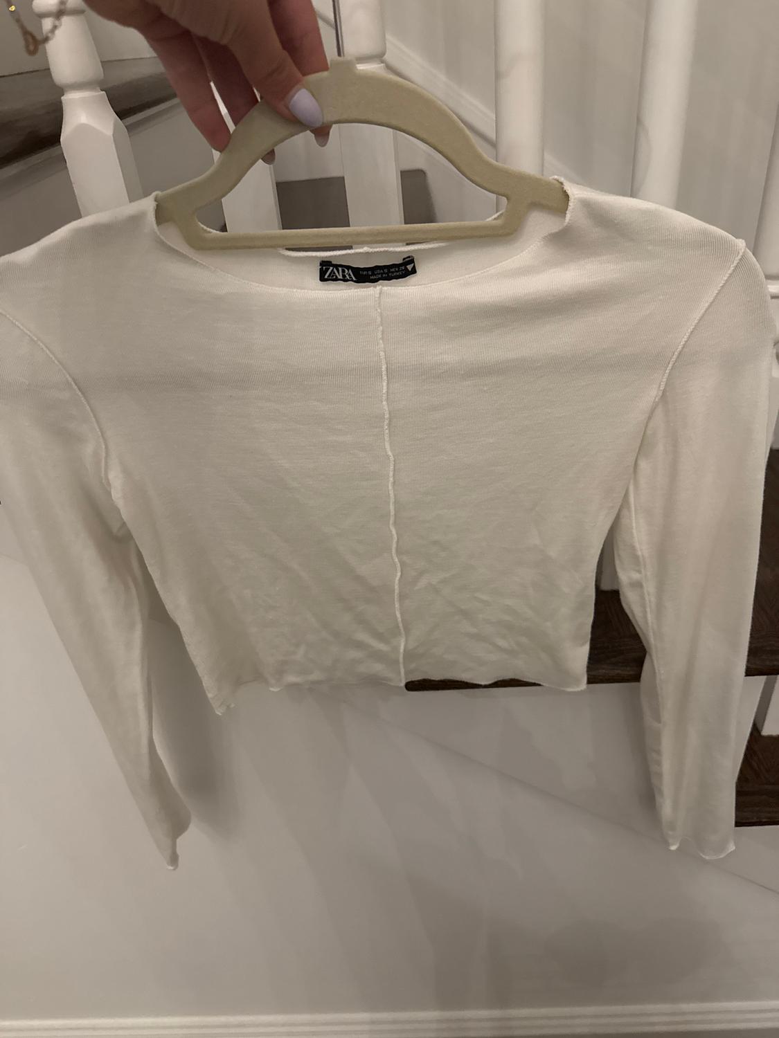 A white, long-sleeved jumper hanging on a hanger, held by a hand with white painted nails. In the background, white stairs are visible.