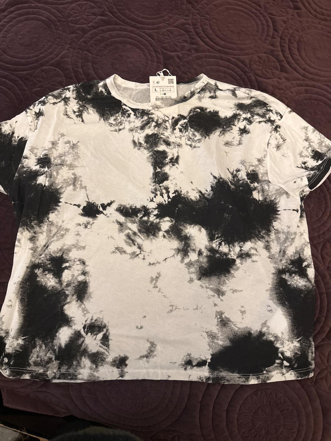 White t-shirt with abstract black print on purple quilt. Visible tag with size L and QR code.