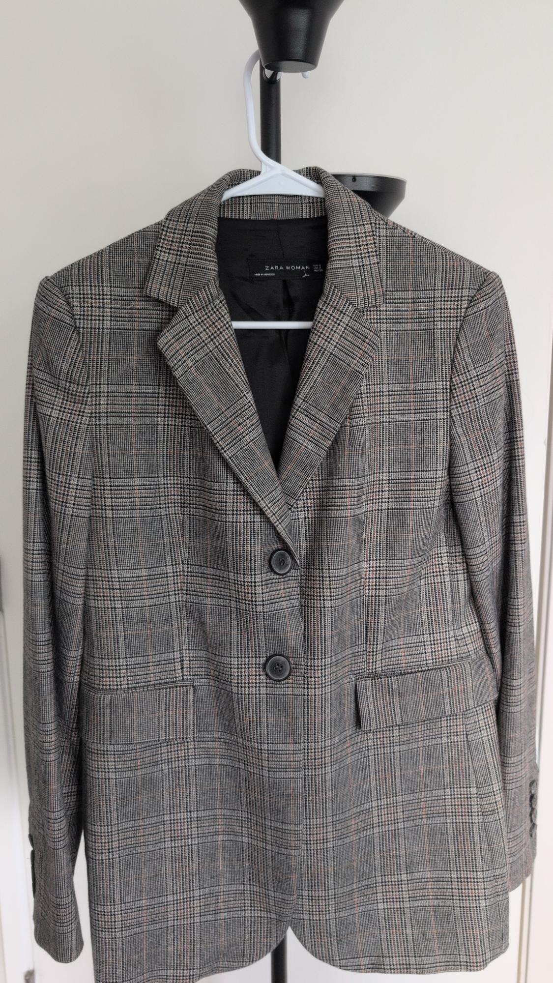 Grey plaid blazer hanging on a hanger. Two-button closure and flap pockets. Interior label with the text ZARA WOMAN.