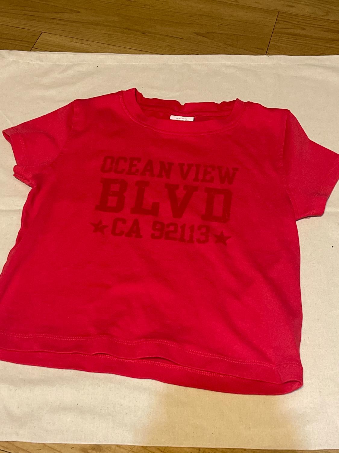 Red T-shirt with text "OCEAN VIEW BLVD CA 92113" on the front, on a light wooden surface and white fabric.