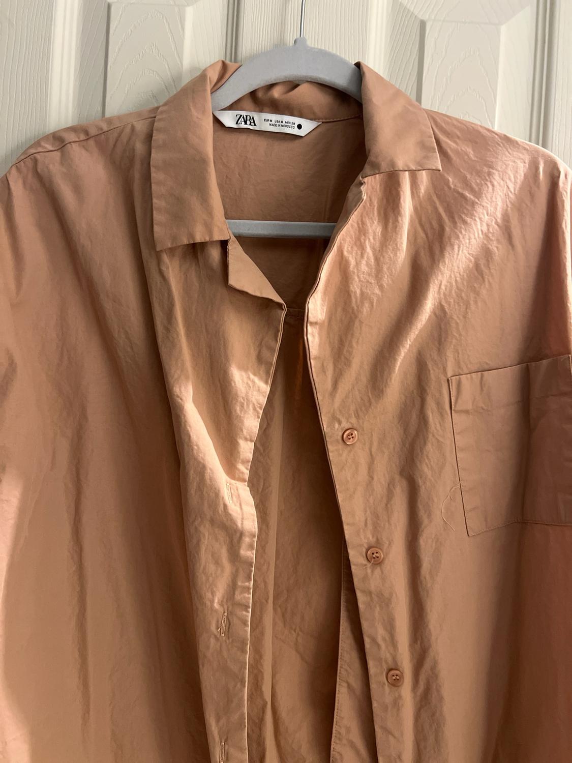 Casual salmon-colored shirt with a pocket, hanging on a white door. Text: ZARA EUR M USA M MEX 28 MADE IN MOROCCO.