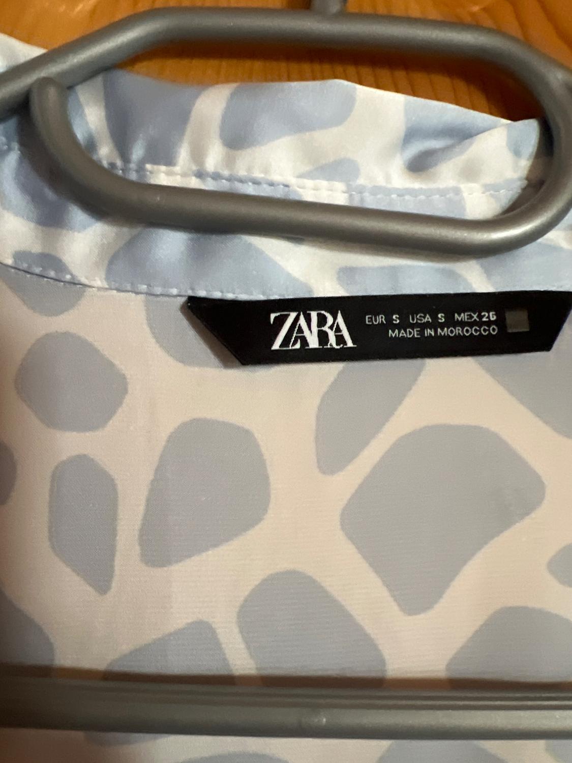 Close-up of a white garment with a light blue print on a hanger. Label: ZARA EUR S USA S MEX 26 MADE IN MOROCCO.