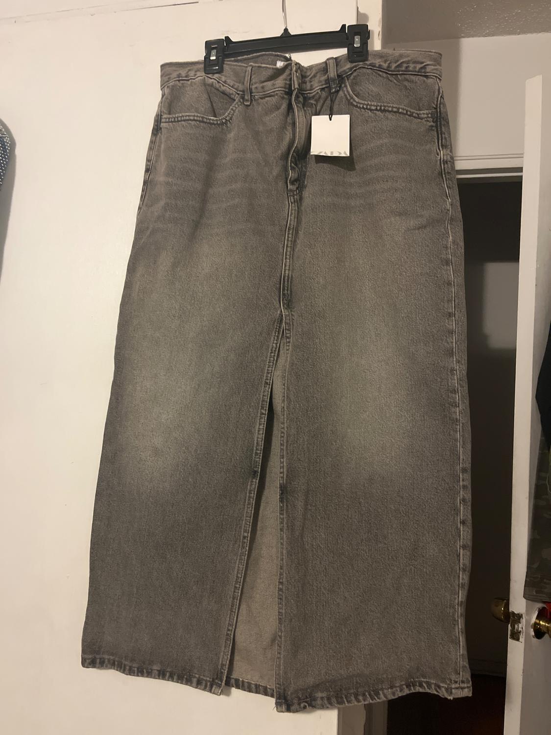 A long grey denim skirt hanging on a black hanger, with a visible white label. White wall background and ajar door.