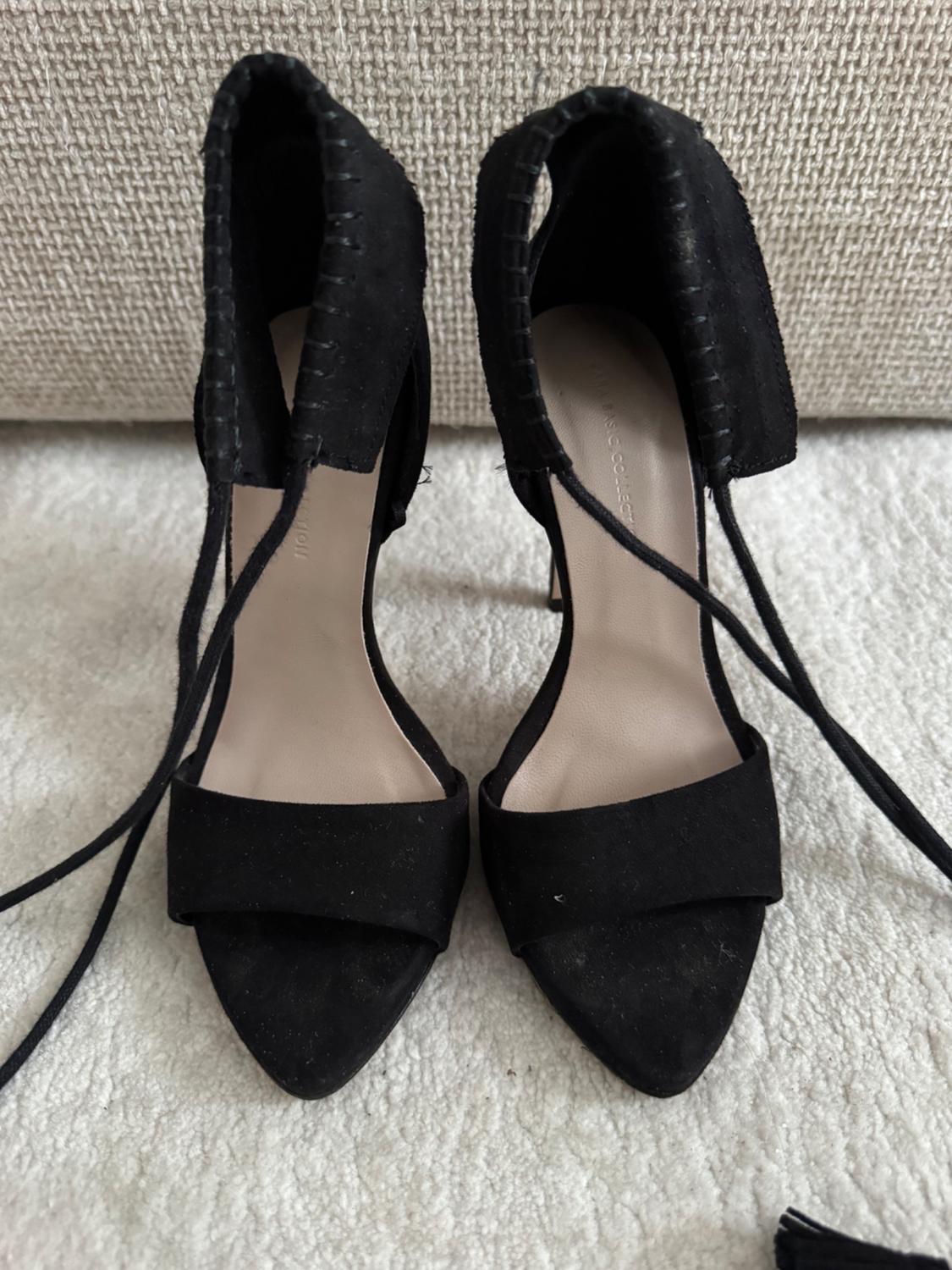 Black high heels with long straps on a white rug, beige fabric background.