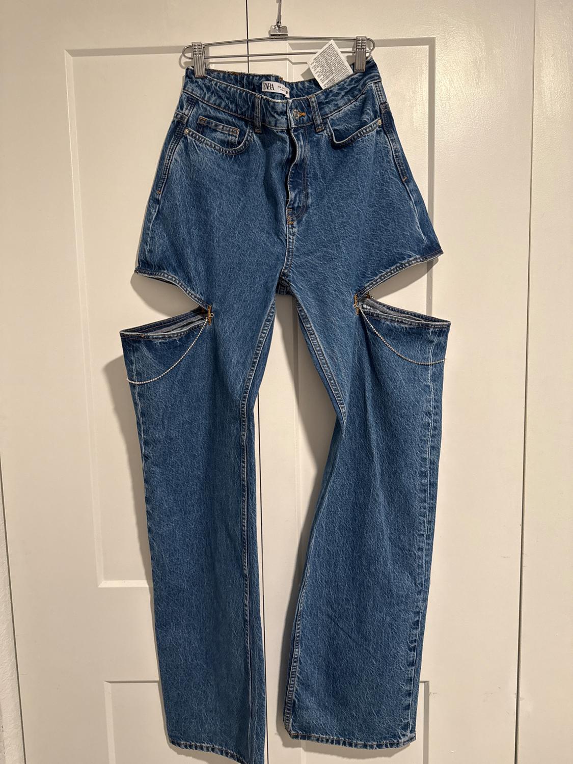 Blue denim trousers with side cuts and zips, hanging on a hanger in front of a white door.