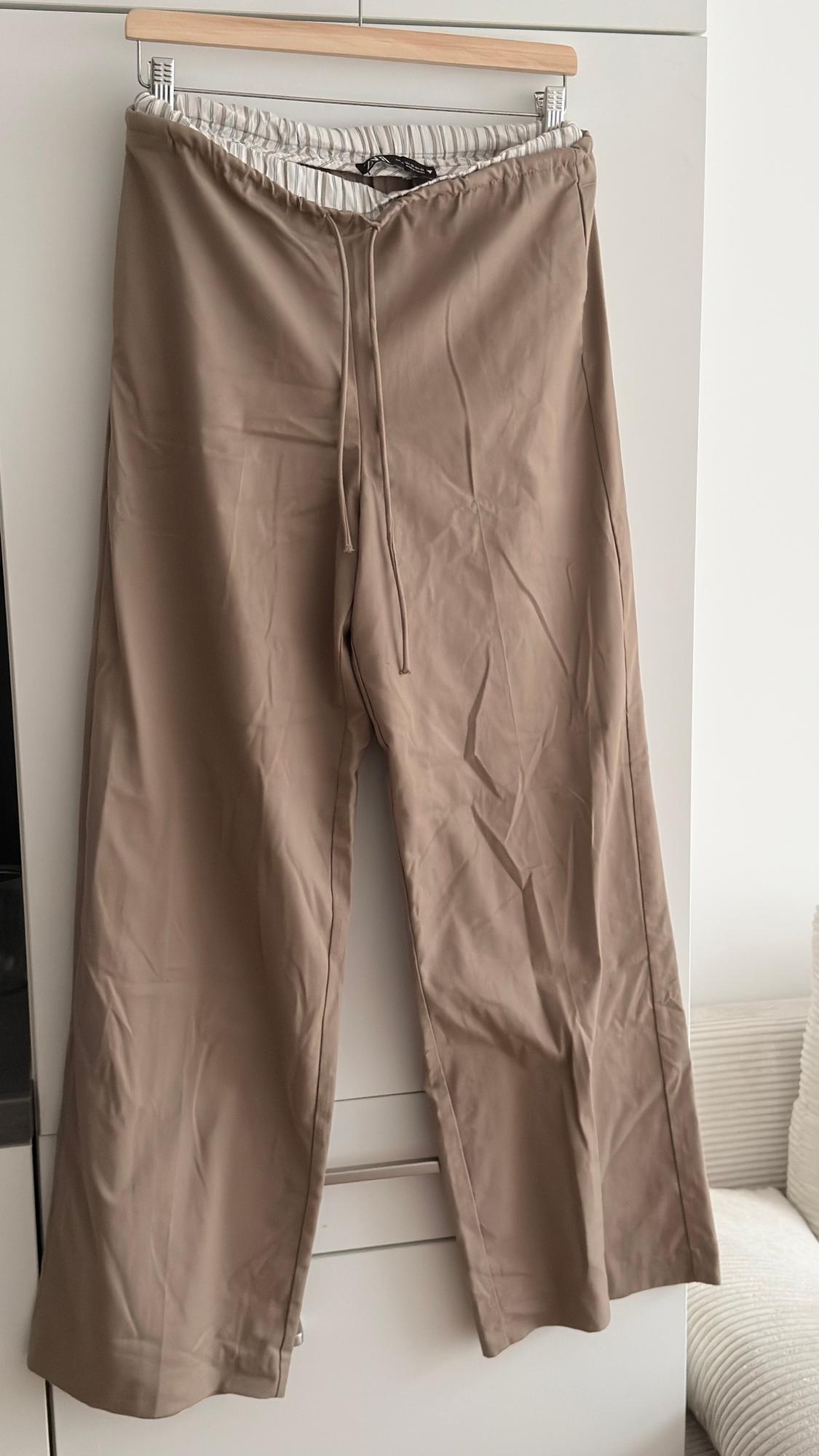 Lightweight beige trousers, made from fabric, hanging on a wooden hanger, with an elasticated waist and adjustable drawstring, against a white background.