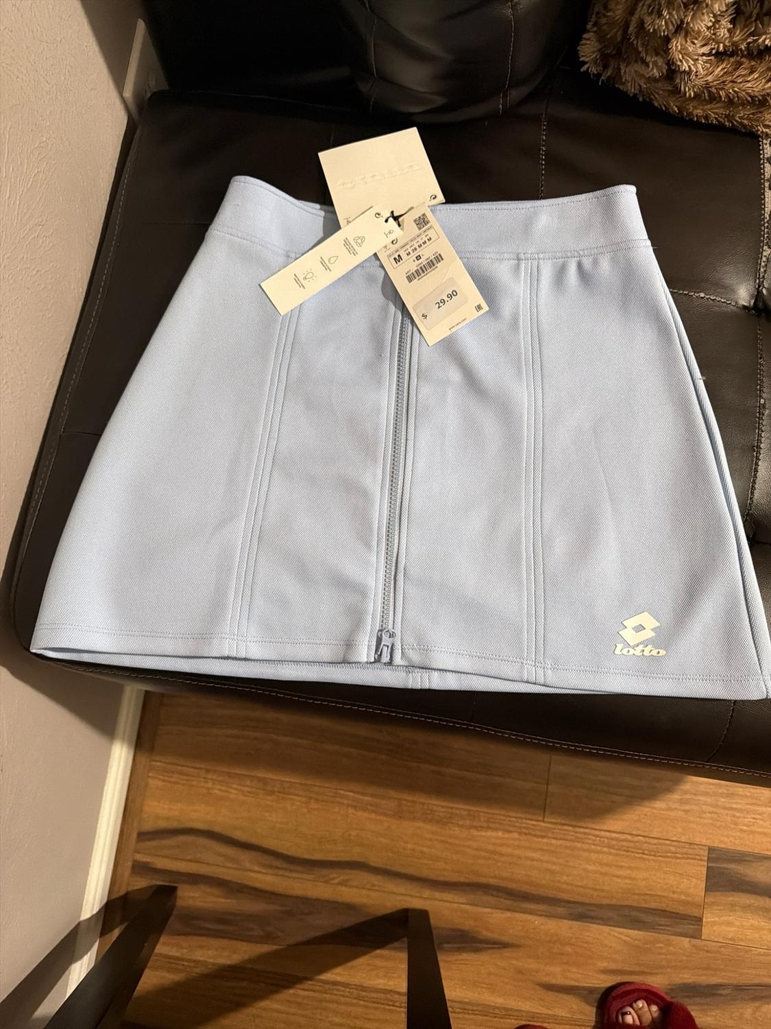 Light blue skirt with front zipper and Lotto logo. It has tags with M and $ 29.90. It is on dark leather and a wooden floor.
