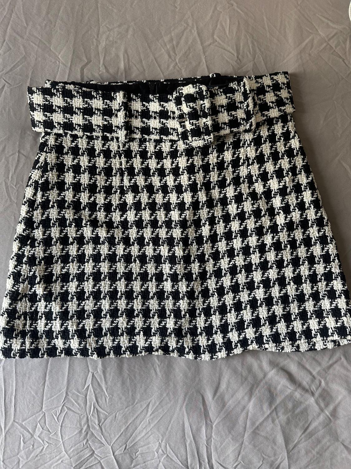 Black and white houndstooth mini skirt with matching belt, on a wrinkled grey fabric background. Relaxed composition.