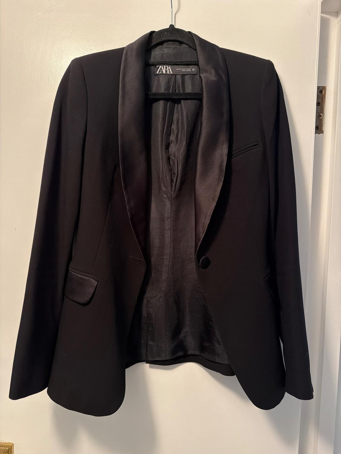 Black satin jacket hanging on a hanger, with wide lapels and a visible Zara label inside. White background.