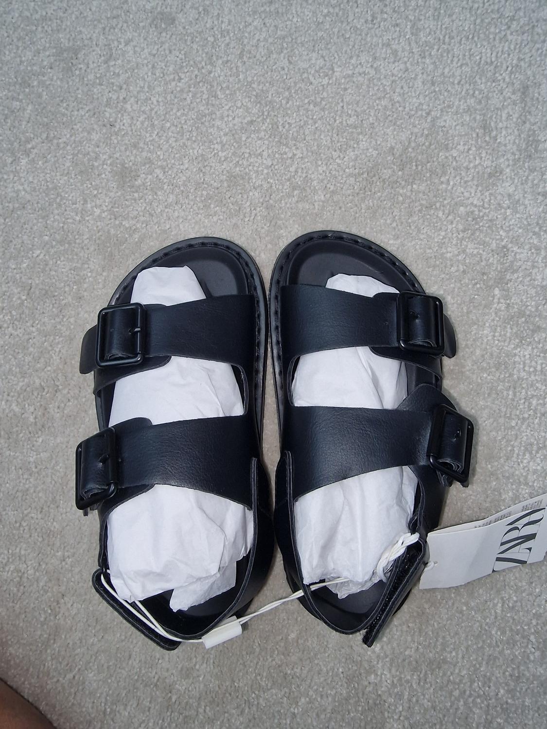 Black two-strap buckle sandals on grey carpet. Visible label with text "ZARA".