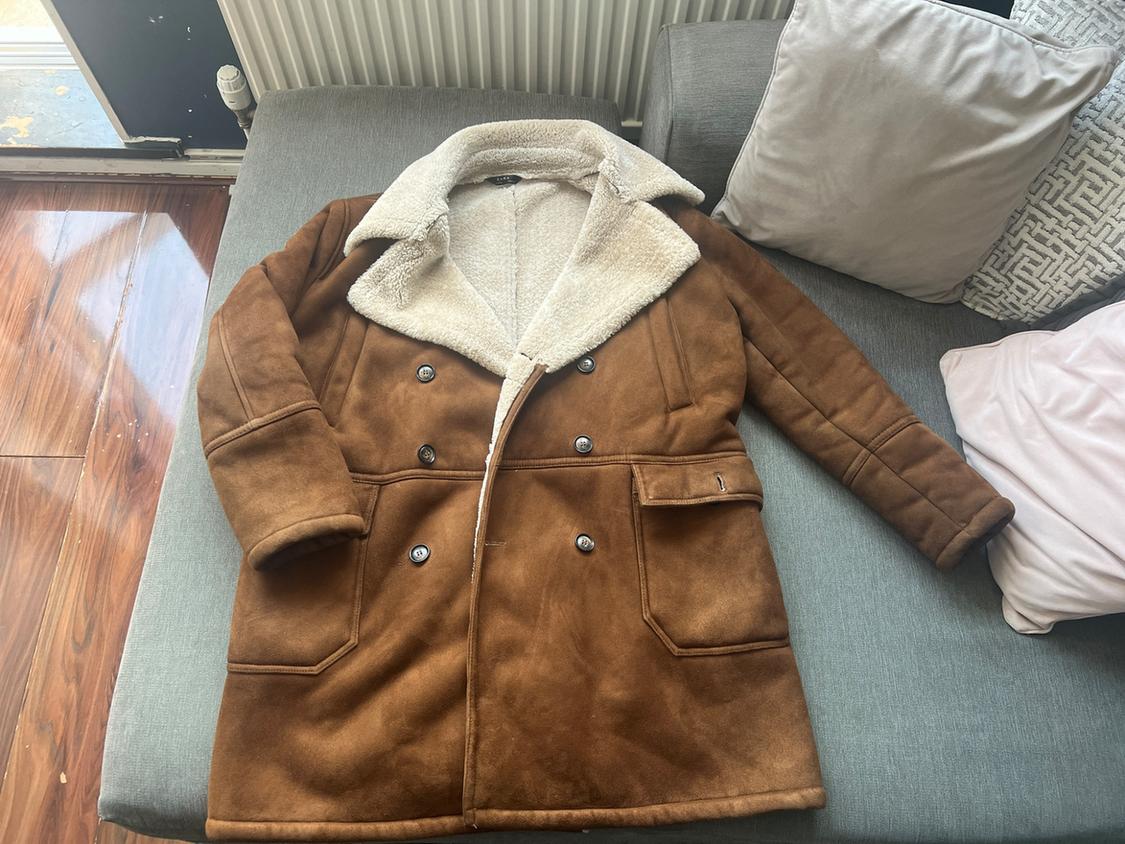 Brown double-breasted coat with a white faux-fur collar and lining, dark buttons and pockets, draped over a grey sofa with cushions.