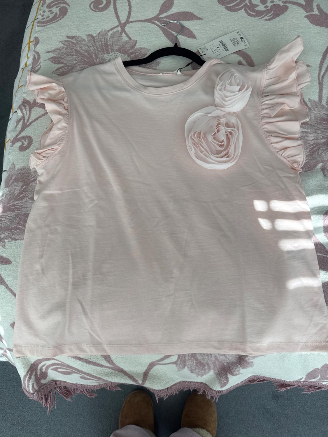 Light pink T-shirt with ruffled sleeves and decorative flowers on the chest, on a bed with a floral bedspread.