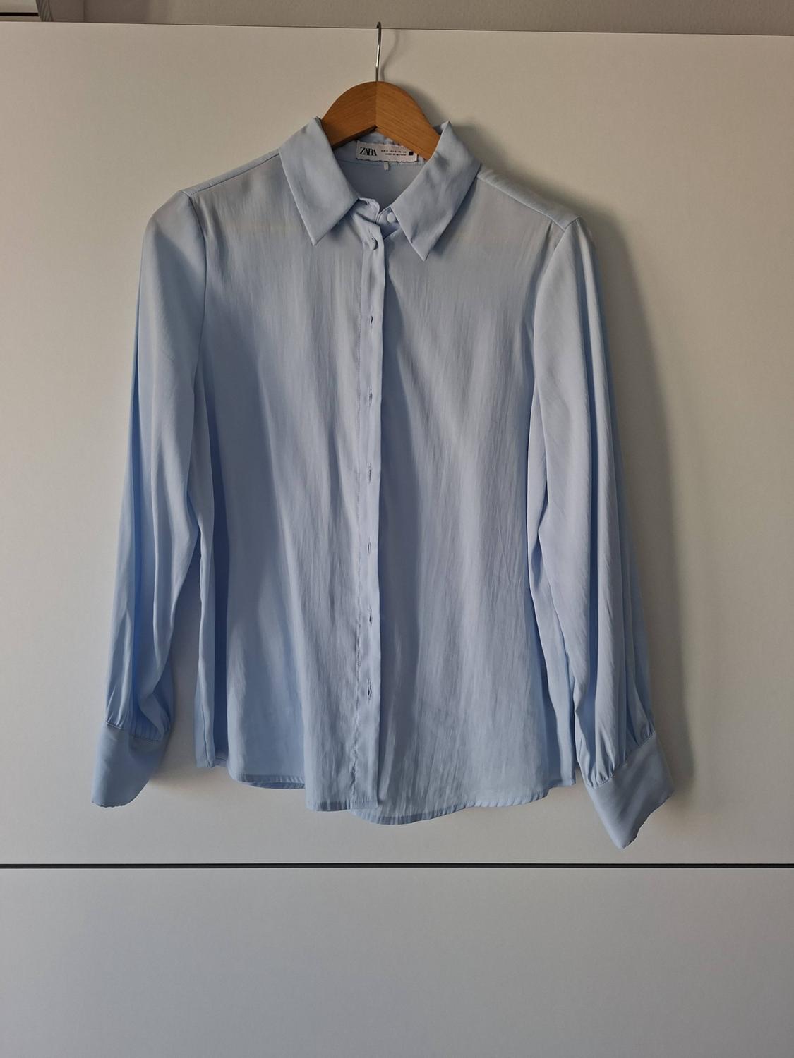 Light blue long-sleeved button-up shirt hanging on a wooden hanger against a white background. The label reads ZARA.