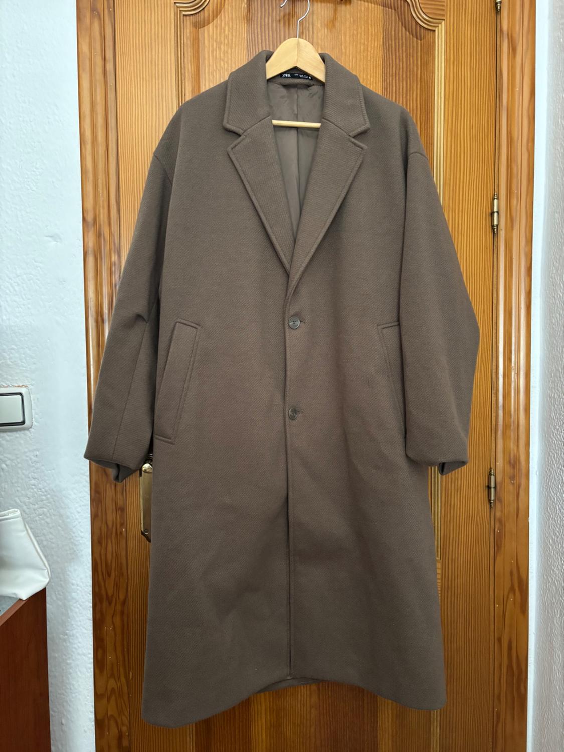 Long brown coat hanging on a wooden door. It has wide lapels and two side pockets. Visible label: "Zara".