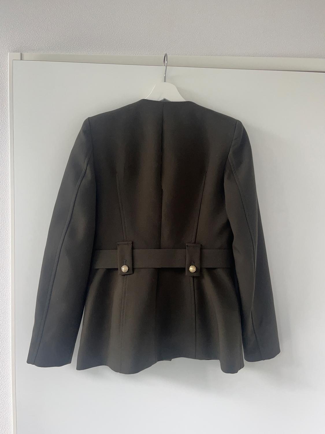 Black jacket hanging on a white hanger against a light background. The back has two decorative buttons at the waist.