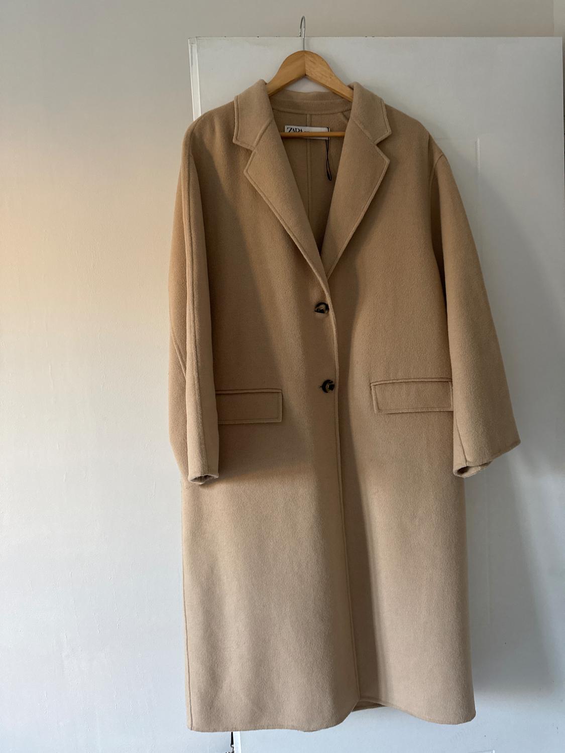 Long beige coat hanging on a wooden hanger over a white door. Visible label with text "ZARA".