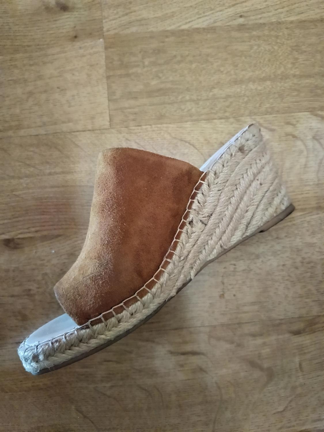 Espadrille-style wedge sandal with a braided jute wedge and brown suede upper, resting on a light wood floor in a casual setting.
