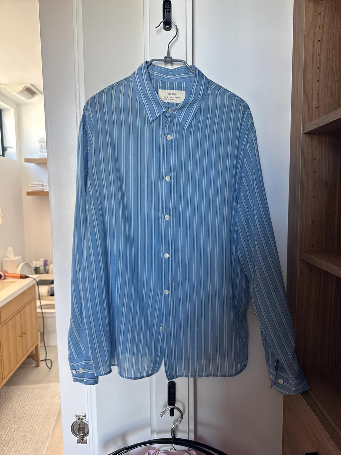Blue and white striped shirt with a ZARA ORIGINS label hanging on a white door. In the background, a bathroom and a wooden shelving unit can be seen.