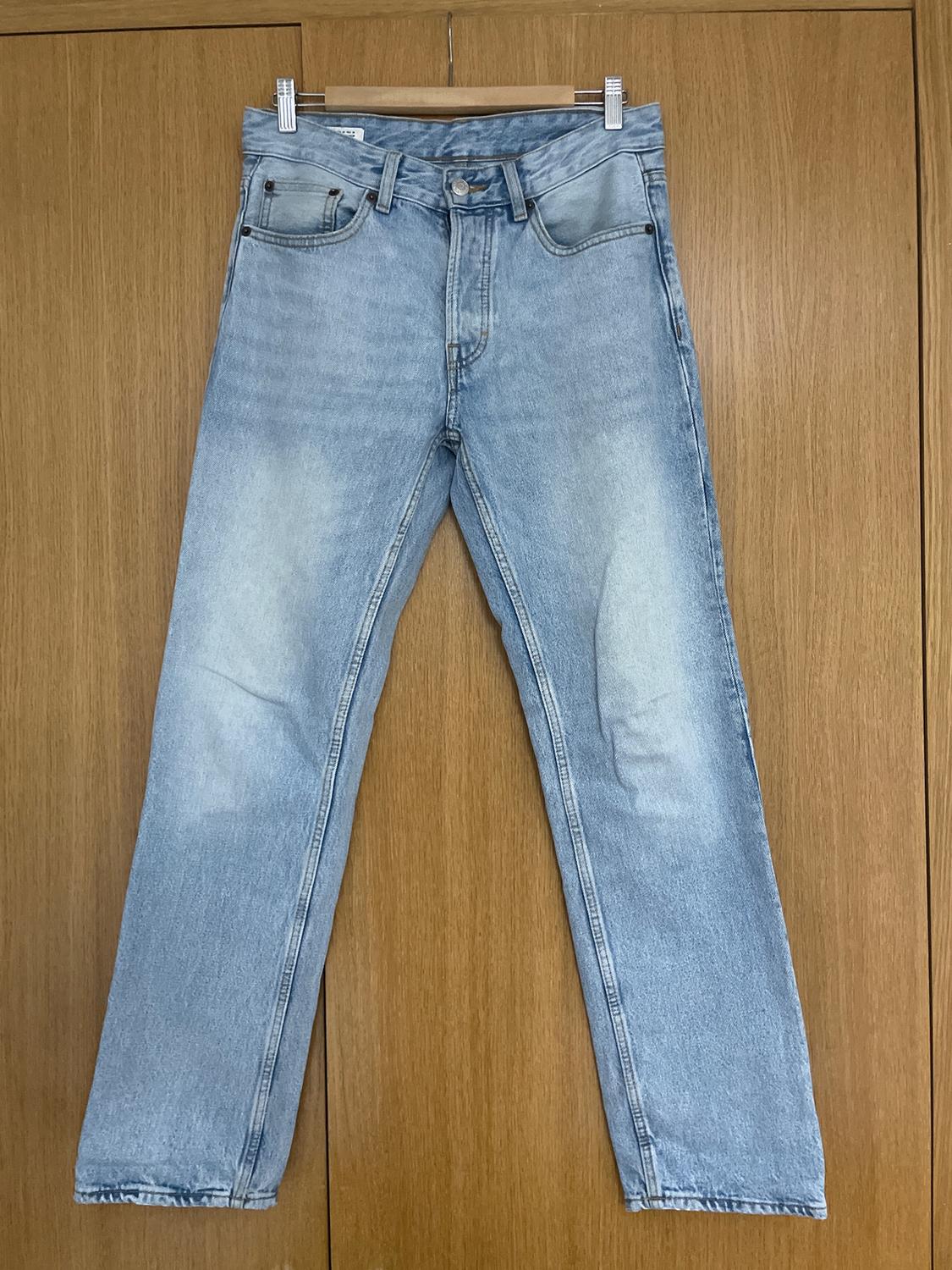 Light blue jeans hanging on a wooden hanger in front of a wooden door. Classic five-pocket design with a button fastening.