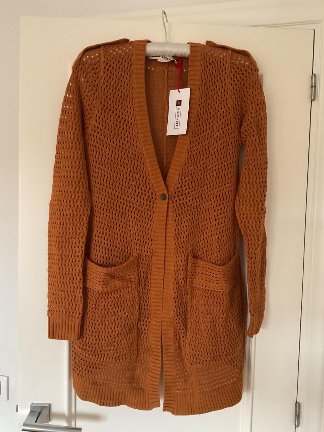Brown knitted cardigan with large pockets, hanging on a white hanger. Visible label with text "Zara Knit". White door in the background.