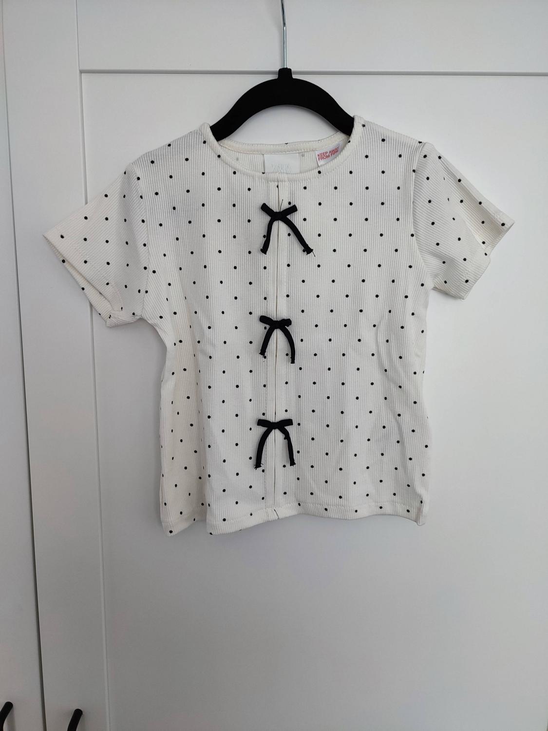 White short-sleeved t-shirt with black polka dots and three bows on the front, hanging on a hanger against a white background. Text: KEEP AWAY FROM FIRE