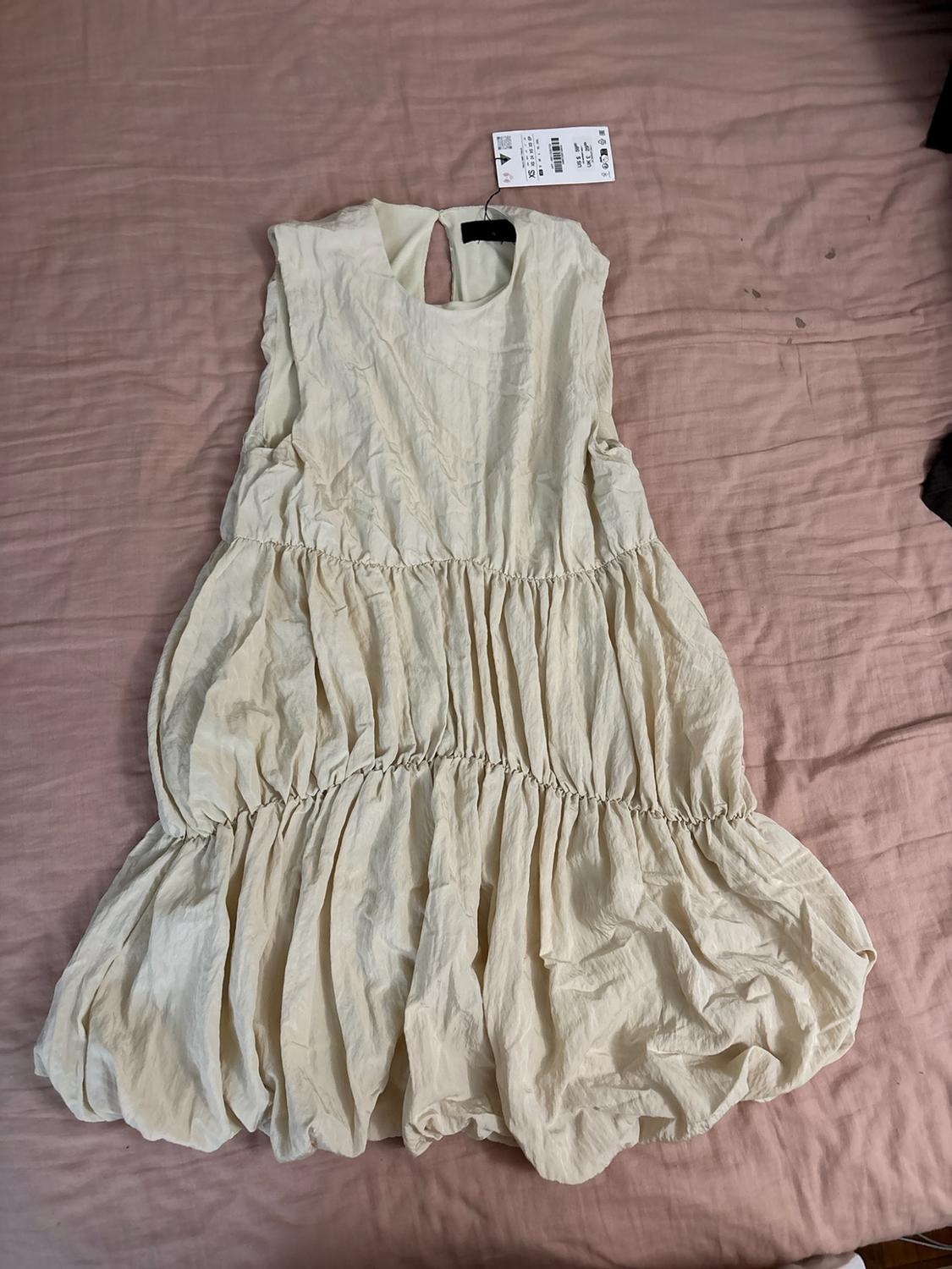 Sleeveless cream-coloured dress with a voluminous gathered tiered design, laid out on pink fabric. Label visible at the neckline.