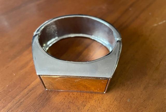 Metallic bracelet with a geometric design on a wooden surface. The bracelet has a shiny finish and a rectangular front panel.