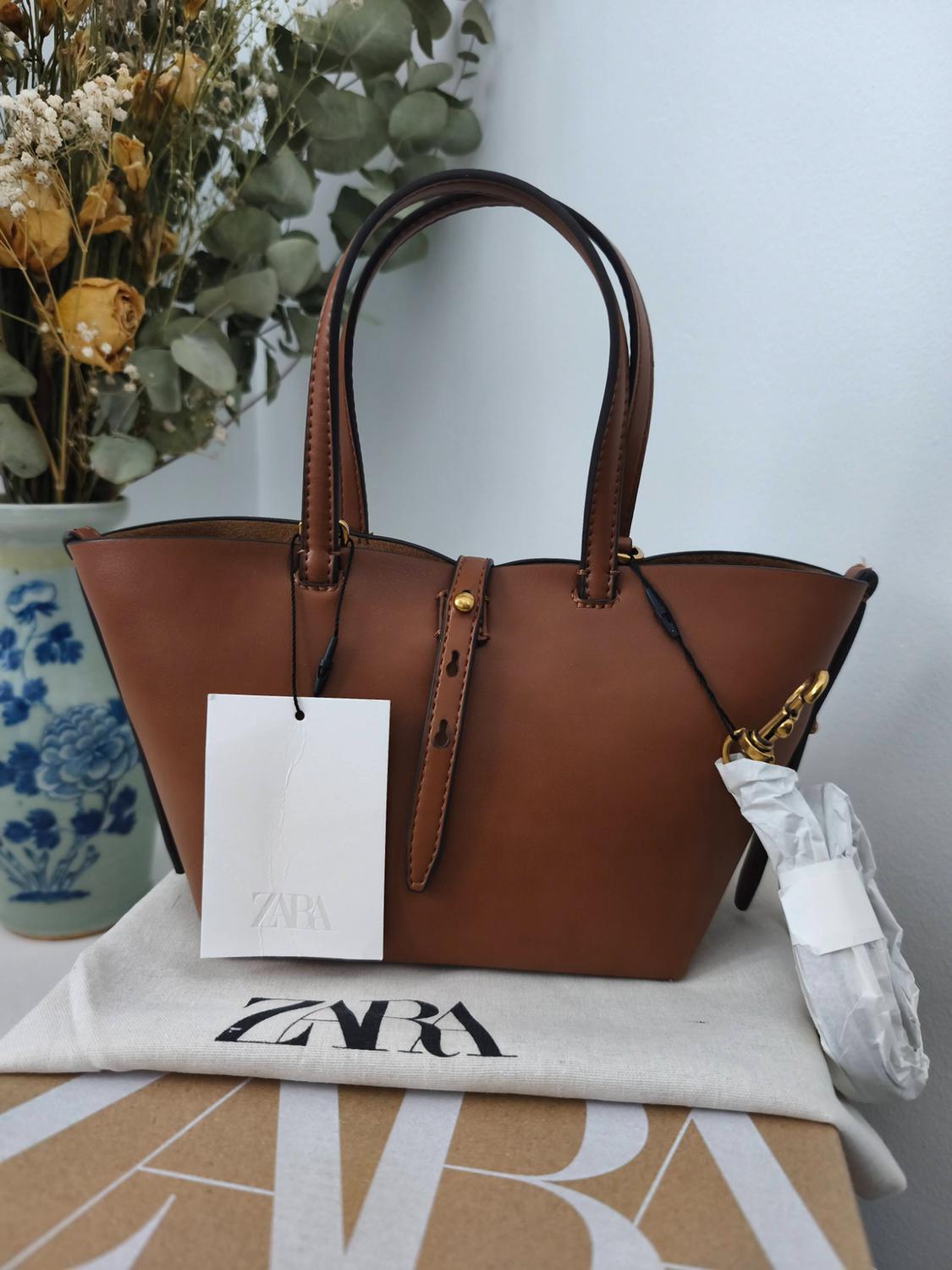 Brown handbag with handles and a ZARA label, on a cover and cardboard box bearing the text ZARA. In the background, a vase with dried flowers.