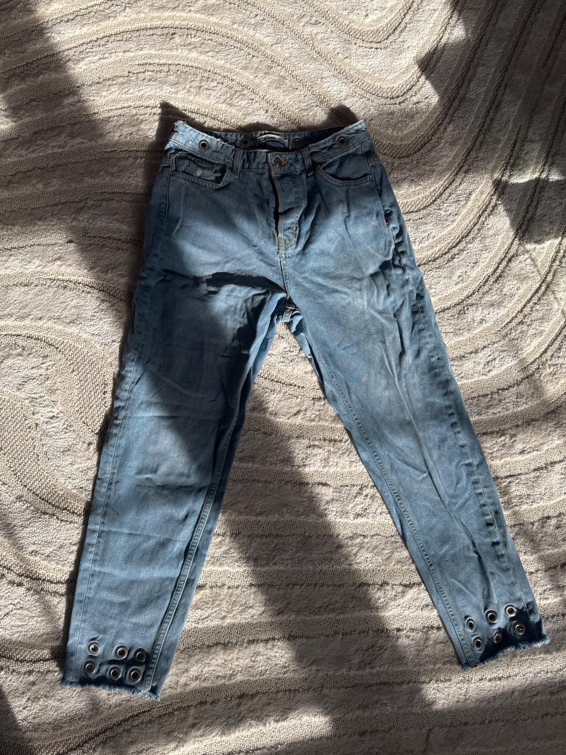 Front view of blue denim jeans with side pockets and eyelets at the ankles.