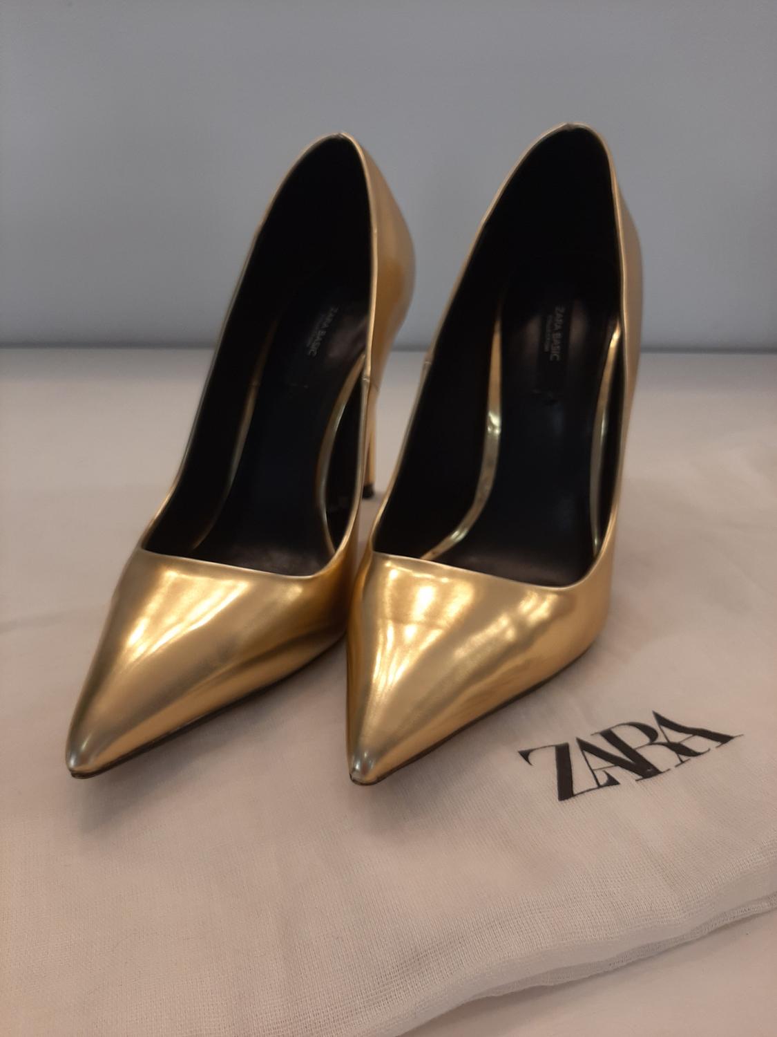 Gold stiletto heels with pointed toe on a white bag with 'ZARA' text.