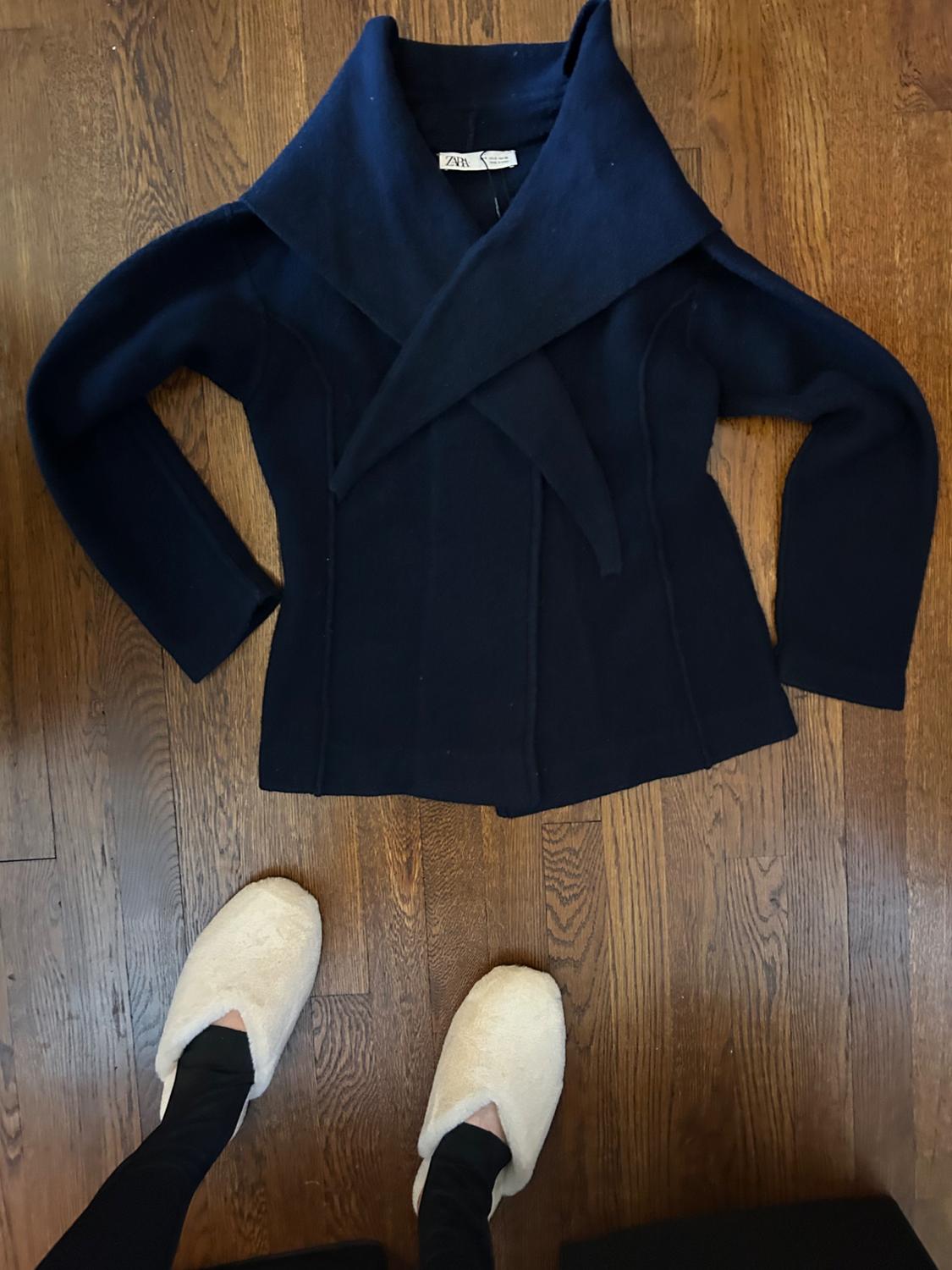 Dark blue coat with a wide collar on a wooden floor. Feet in white slippers visible at the bottom of the image.