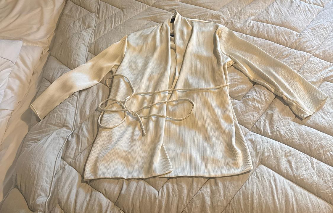 Long-sleeved white textured blouse with a tie belt, laid out on a light-coloured duvet on a bed in a casual and relaxed style.