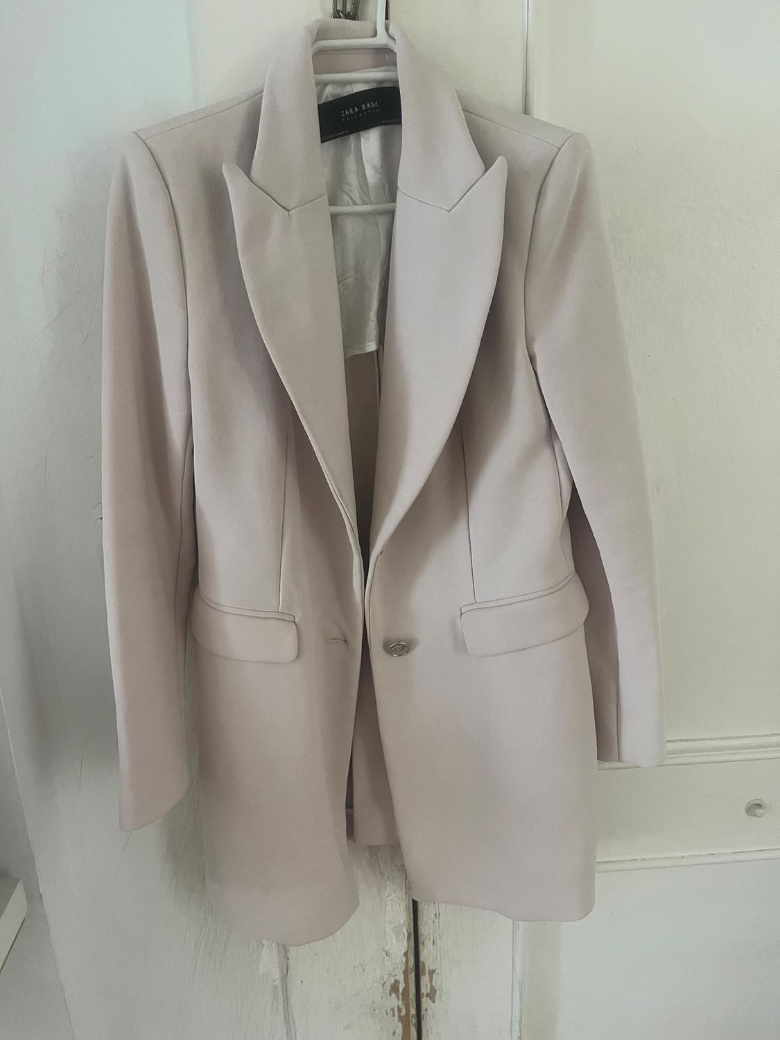 Beige blazer hanging on a hanger, with wide lapels and front pockets. Visible label: "ZARA BASIC". White background.